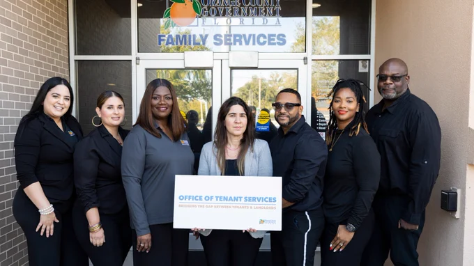 A group of seven people stands outside the Orange County Government Family Services building. They hold a sign that reads "Office of Tenant Services," promoting support in various languages. The group is dressed in professional attire.