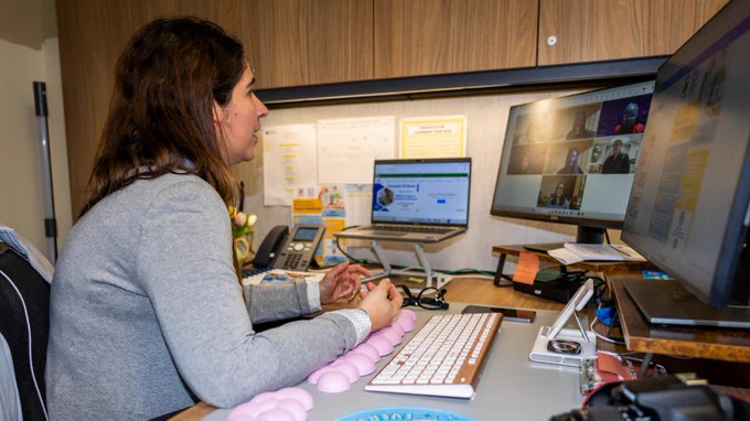 A person sits at a desk in an office, engaged in a video conference call on two computer screens. Several people appear on the screens. The desk has a computer keyboard, a camera, and papers.