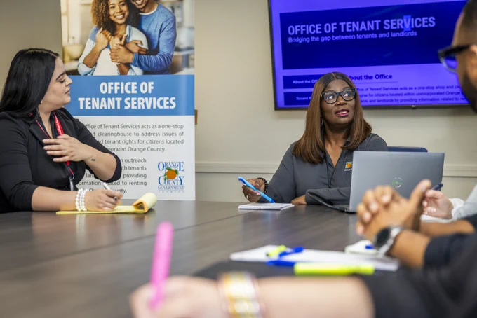 A group of people sit around a conference table engaged in discussion. A sign reads "Office of Tenant Services," and the background screen displays information about tenant services. Various office supplies are on the table.