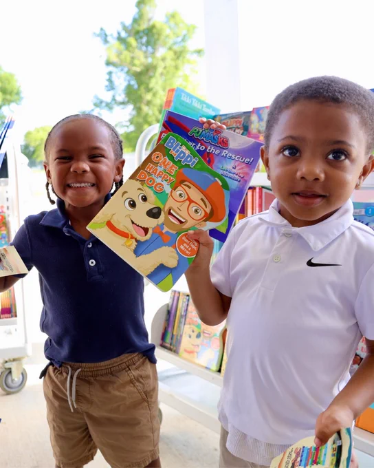 Two young boys, one in a navy polo shirt and the other in a white t-shirt, standing in a library or bookstore and holding a colorful children's book.