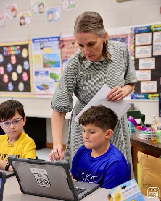 A teacher assisting two students working on a laptop in a classroom setting.