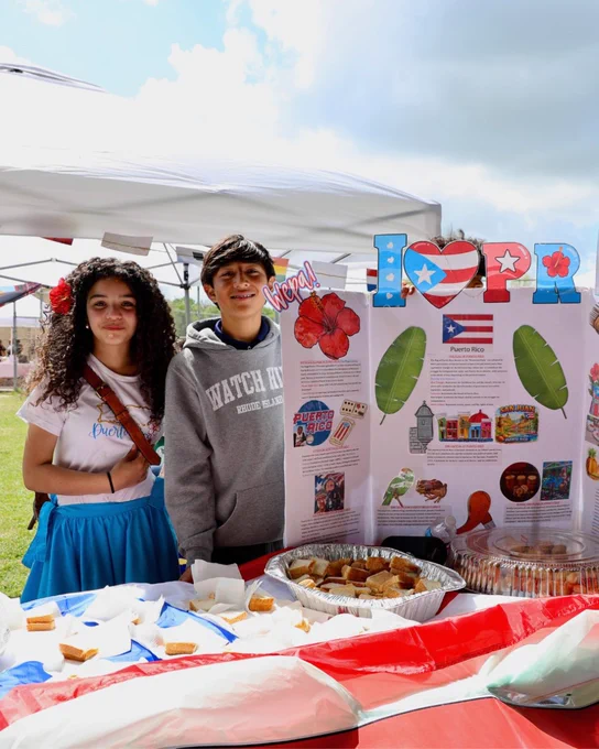 Two young people stand in front of a colorful festival booth displaying information about Puerto Rican culture and cuisine.
