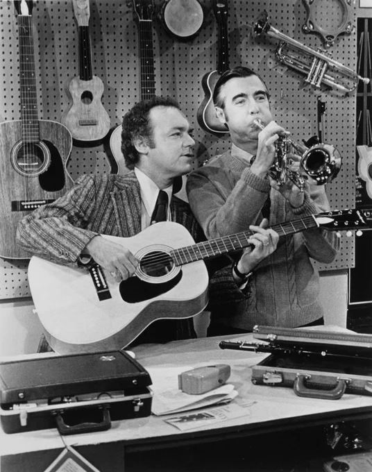 Joe Negri playing a guitar and Fred Rogers playing a trumpet in the Negri Music Shop surrounded by hanging string instruments and instrument cases on the counter.