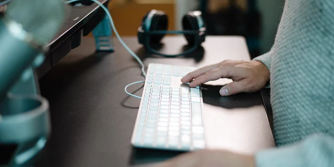 A person wearing a sweater is typing on a keyboard at a desk. Nearby, there is a microphone and a pair of headphones resting on the surface.