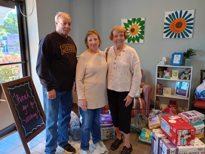 Three adults standing inside a room with baby supplies such as diapers and wipes and colorful flower paintings on the wall behind them.