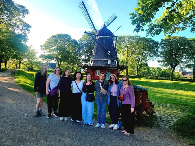 A group of girls posing near a historic windmill in a sunny park with green trees and a blue sky.