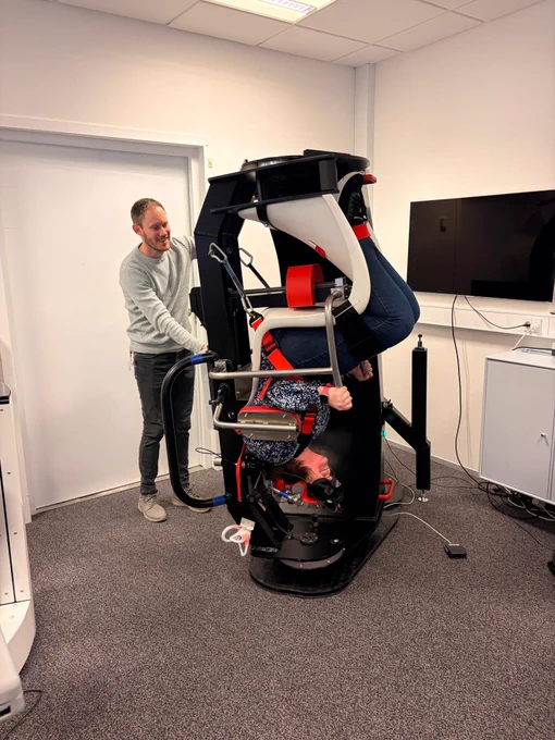A person is secured upside down in a medical or scientific testing device while another person stands nearby, monitoring the process in a clinical room.