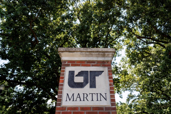 A brick campus sign displaying the UT Martin logo stands beneath a canopy of large, leafy trees.