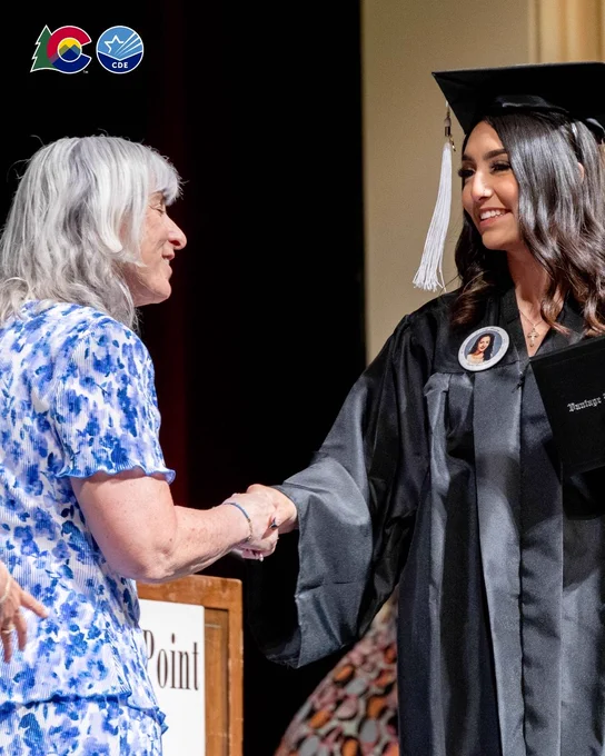 A person in a floral outfit is shaking hands with a graduate wearing a black cap and gown. The graduate is holding a diploma. The Colorado State and CDE logo are visible above them.
