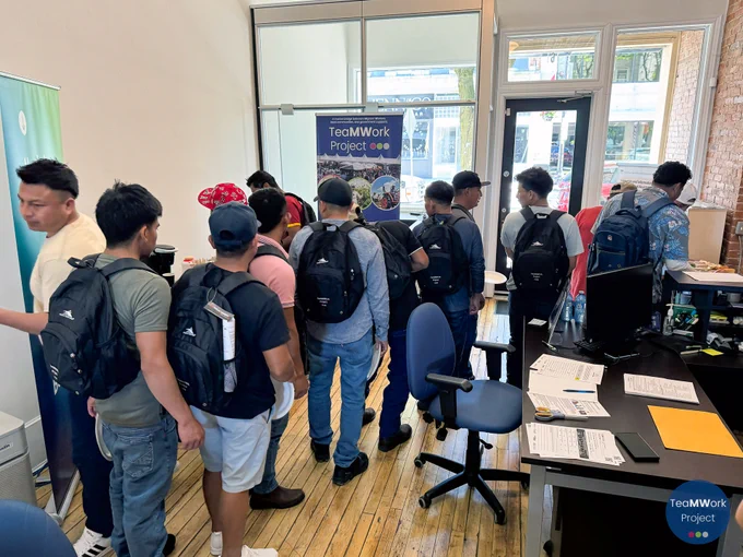 Temporary foreign workers line up inside Collège Boréal’s Chatham-Kent office to receive refreshments and materials, standing near a TeaMWork Project banner during a workplace eye safety workshop.