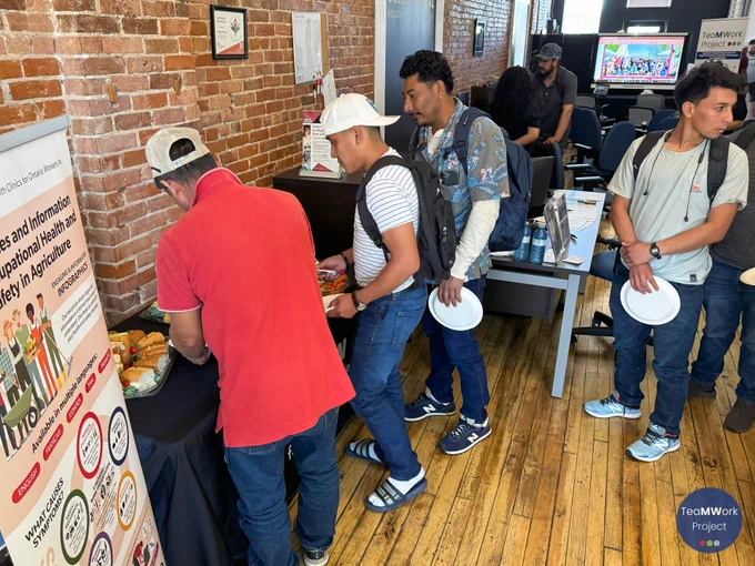 Temporary foreign workers select food from a refreshment table inside the workshop space, with informational posters about occupational health and safety visible beside them.
