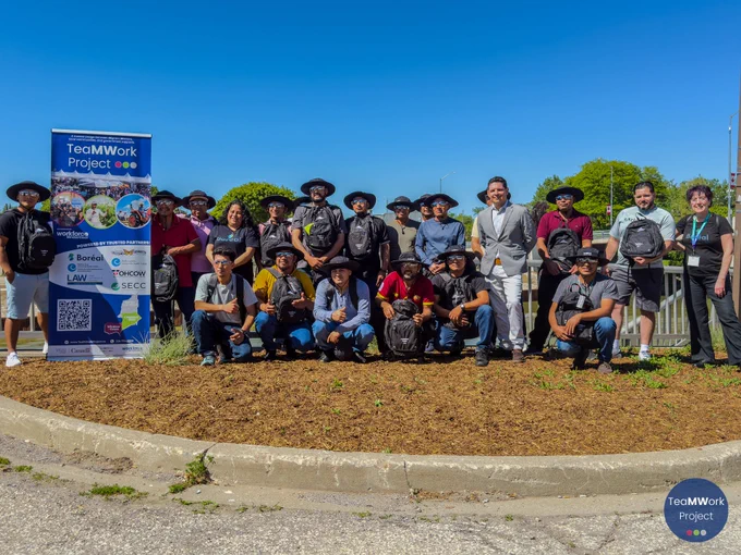 Group photo outdoors of temporary foreign workers and organizers gathered beside a TeaMWork Project banner on a sunny day, many holding backpacks and items received during the eye safety workshop.
