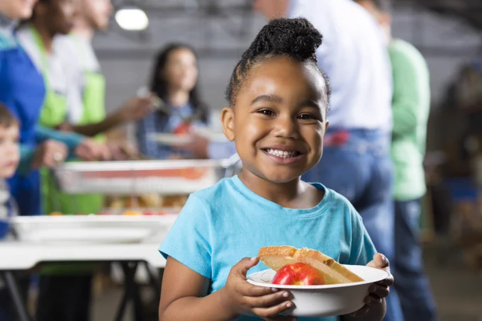 A smiling child holding a bowl with an apple and bread, standing in a room with people serving food in the background.