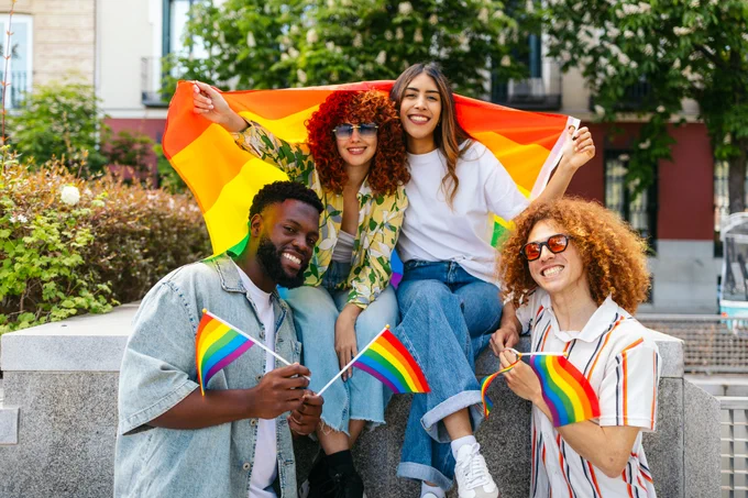 A group of four people joyfully pose outdoors with a large rainbow flag and three small rainbow flags. They are sitting on a low wall in a sunny setting with greenery and buildings in the background.