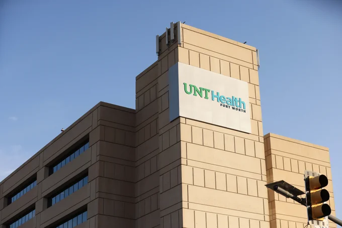 The image shows a building with a sign that reads "UNT Health Fort Worth" against a clear sky. There is a traffic light in the foreground.
