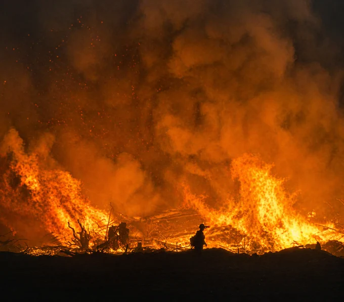 A firefighter silhouetted against towering flames and thick smoke during a large prescribed burn at night. 
