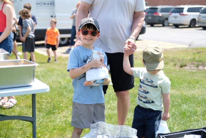 Two young boys holding hands with an adult while standing on grass near a table and parked cars on a sunny day.