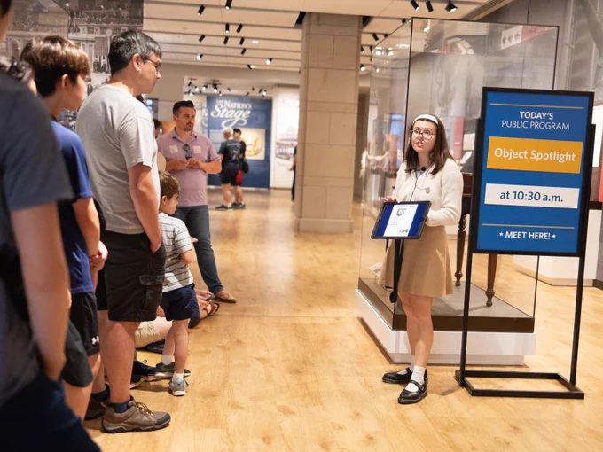 A person is presenting to a small group inside a museum. They are holding a tablet and standing near a sign that reads, "Today's Public Program: Object Spotlight at 10:30 a.m." In the background, there are museum displays and a sign with "Nation's Stage.
