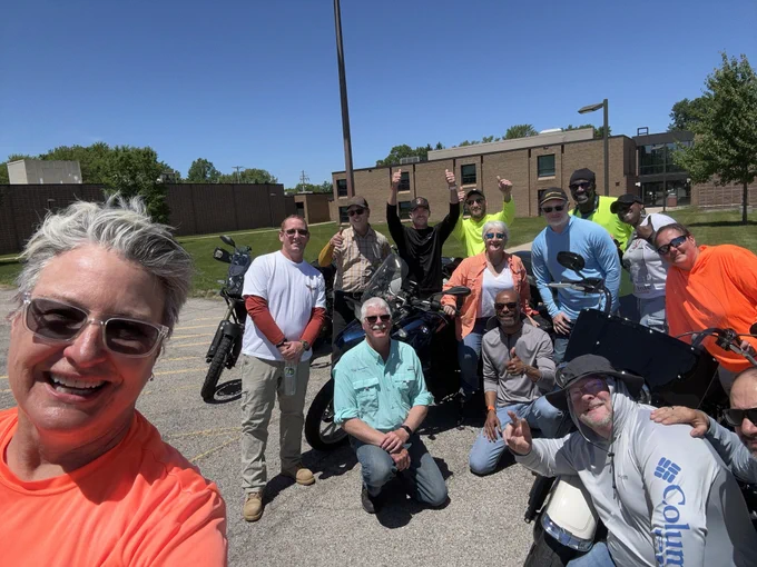 A group of people posing in a parking lot with motorcycles. They are smiling at the camera, and some are wearing sunglasses and casual outdoor clothing. The background features a brick building and a clear blue sky.