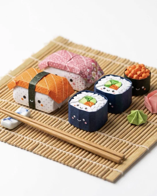 Origami sushi pieces arranged on a bamboo mat with chopsticks. Cute paper sushi art includes nigiri, maki, and ikura with kawaii-style faces, blending traditional cuisine with playful design.