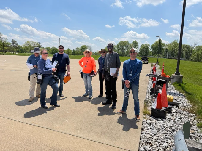 A group of people stands on a paved area outdoors on a sunny day, with a row of orange traffic cones nearby. The background features green trees and a blue sky with clouds.