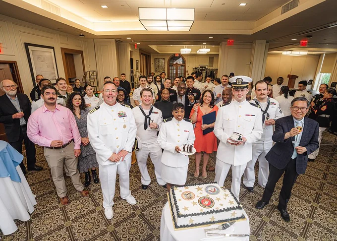 Dozens of Columbia University students and faculty as well as guests, either dressed in white naval uniforms or suits or dresses smile and raise their glasses and slices of ceremonial cake at the NROTC commissioning ceremony at Columbia University Faculty House.