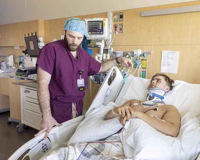 A doctor in maroon scrubs and a blue surgical cap stands beside a hospital bed, talking to a young patient with a neck brace lying under white blankets. Medical equipment and monitors are visible in the background of the hospital room.