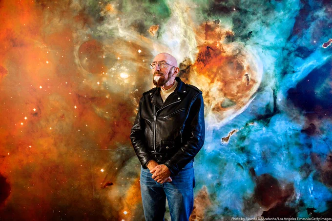 Kip Thorne wearing jeans and leather jacket in the foreground looking upward. In the background is a colourful cosmic scene.