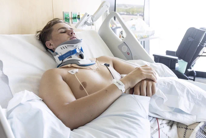 A young man is lying in a hospital bed with a cervical collar supporting his neck. He has medical electrodes attached to his chest and a hospital wristband on his arm. His eyes are closed and his hands are resting on his abdomen. The room is bright, with medical equipment visible in the background.