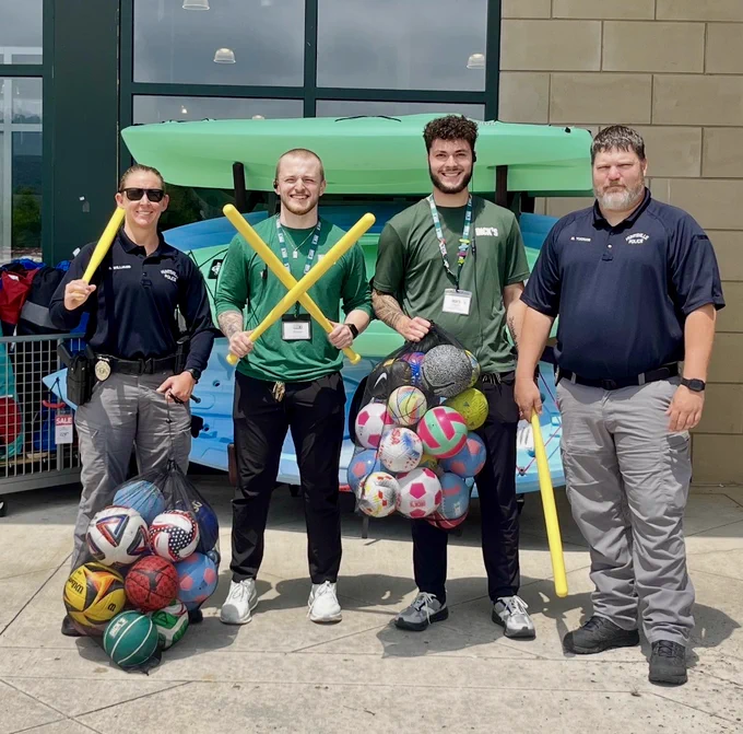 Four individuals are standing outdoors in front of a building, holding sports equipment. Two people in green shirts hold yellow plastic bats, and two others in dark shirts stand on either side. Several mesh bags filled with assorted sports balls, including basketballs, soccer balls, and volleyballs, are on the ground or being held. Behind them are stacked kayaks and store displays.