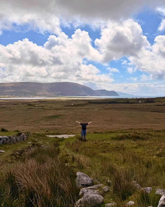 Person standing with arms raised in a vast grassy field under a cloudy sky with distant hills and coastline.