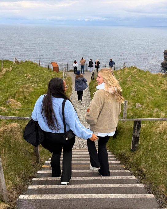 Two people walking down steps toward a coastal viewpoint with grassy hills and a body of water in the background.