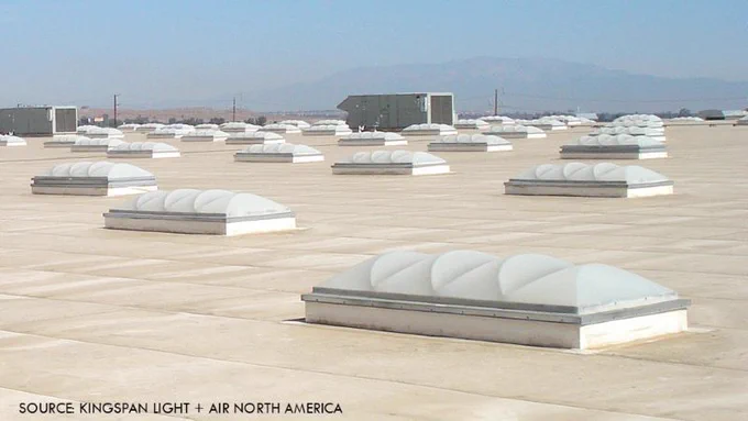 Photo shows flat commercial roof covered with multiple evenly spaced dome skylights, with rooftop equipment and distant mountains visible in the background.