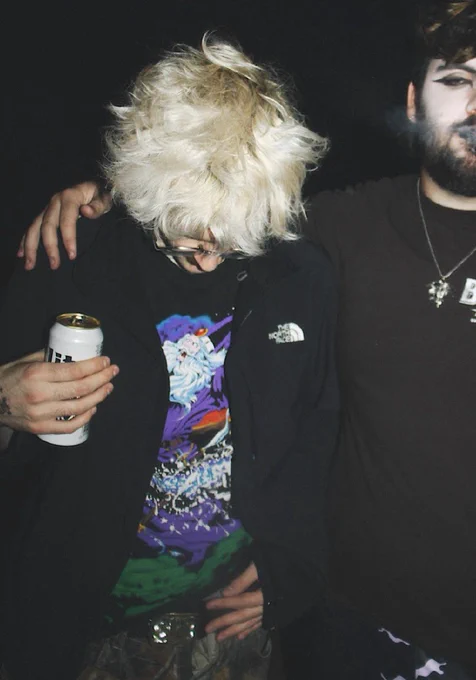 natural blond with a weed leaf belt buckle, north face windbreaker, and a wizard shirt looking at the ground holding a beer