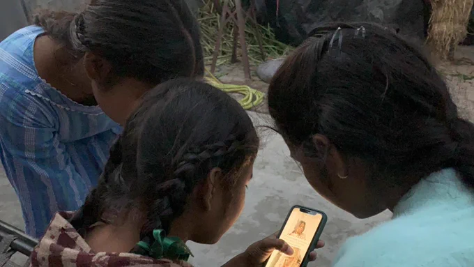 Three young girls in a rural setting gather around a smartphone, closely watching content on the screen together.