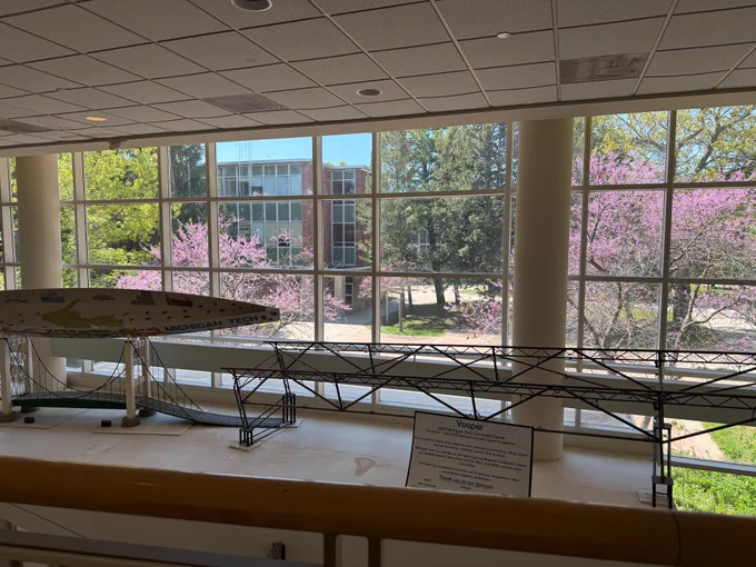 View from inside Michigan Tech’s Dow Building looking out through large floor-to-ceiling windows on a bright spring day. Pink flowering trees and green foliage frame the campus walkway outside. In the foreground, displays of Michigan Tech’s award-winning Concrete Canoe and Steel Bridge projects are showcased inside the building, highlighting student engineering achievements against the colorful spring backdrop.