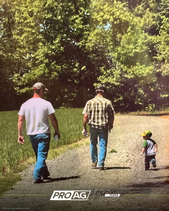 Two adults and young child are walking on a dirt path surrounded by greenery at Spillman Farms. The text on the image reads: "JUNE FEATURE PHOTO - Taken in 2018, this photo captures three generations walking back to the truck after field scouting. It's a meaningful moment of a family working together." The bottom includes logos for ProAg crop insurance company and a 100-year celebration. ProAg is an equal opportunity provider. 