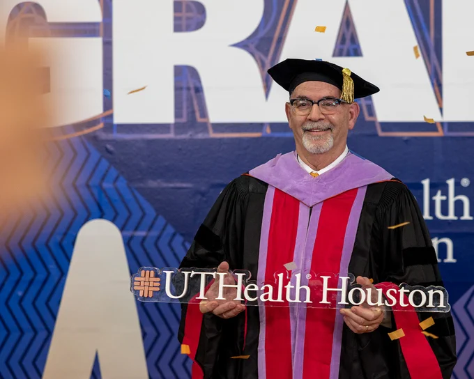 A person wearing academic regalia — a black gown with red and purple accents and a black cap with a gold tassel — stands in front of large letters that partially spell “GRADS.” They hold a sign that reads “UTHealth Houston” while confetti falls around them.