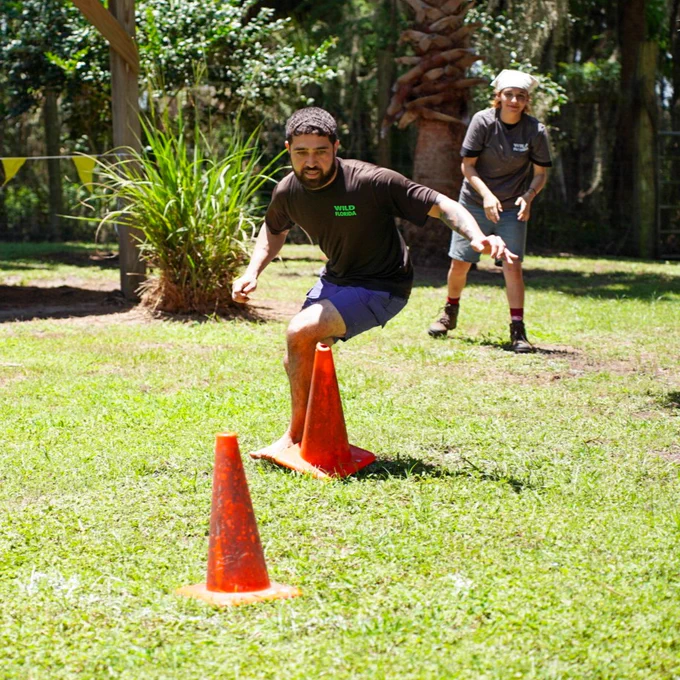 A person in a "Wild Florida" shirt is balancing on an orange traffic cone on grassy ground, with another person observing in the background. Trees and lush vegetation are in the surroundings.