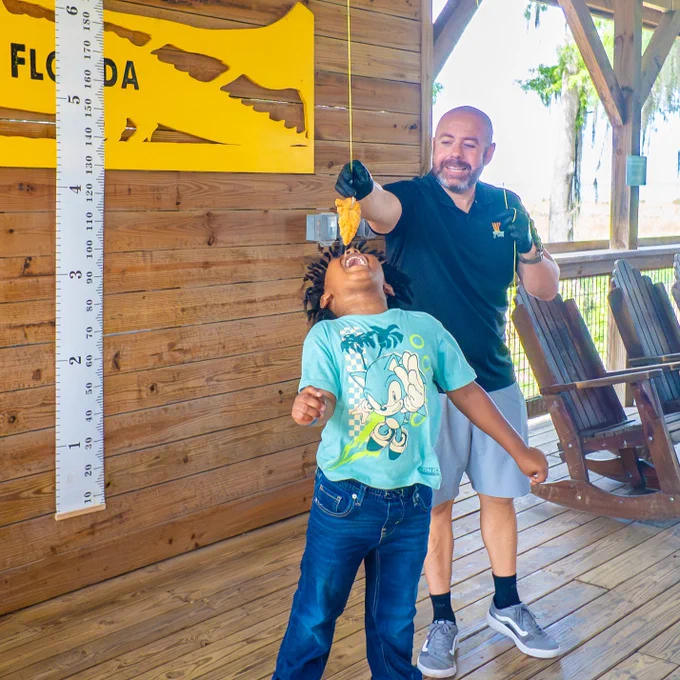 A person playfully holds a piece of food above another individual who is reaching up to bite it. They are in a wooden outdoor setting with a large ruler and part of a yellow sign showing the word "Wild Florida."