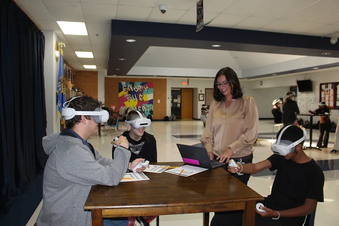 Morgan Dutton assists three students wearing virtual reality headsets at a wooden table in a school commons area.