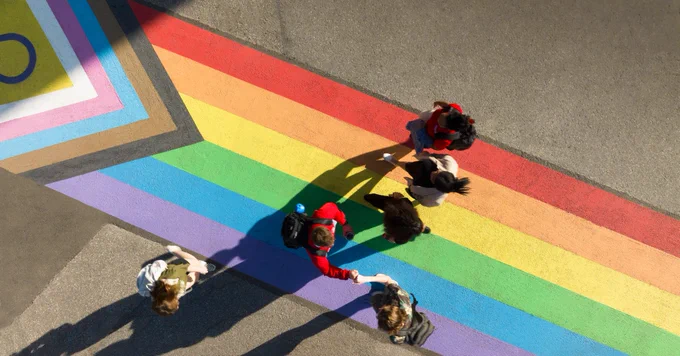 Aerial view of pedestrians crossing a rainbow-painted crosswalk incorporating additional colors symbolizing inclusivity, on a sunny day.
