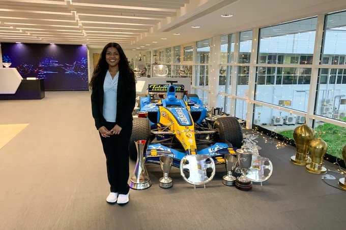 Aarabhi Krishnakumar stands in front of a blue and yellow Formula One race car displayed at the Alpine team office in Oxford, England, with several silver trophies arranged in front of the car. 