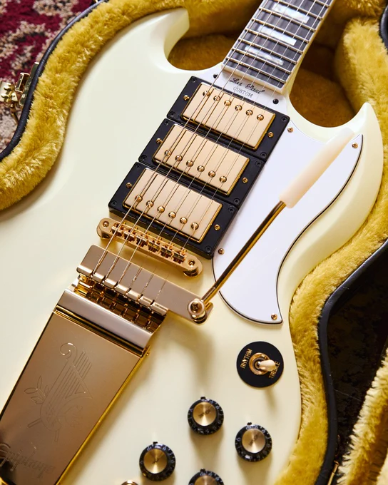 A white Epiphone 1963 Les Paul SG with Maestro Vibrola electric guitar with gold hardware and three pickups is resting in a yellow-lined case. The guitar has a vibrato arm and multiple control knobs.