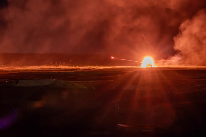 Night live-fire exercise showing a bright explosion and streaking tracer rounds across a dark training area, illuminating the terrain and smoke.