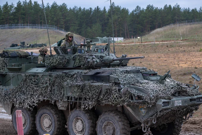 An eight-wheeled armoured vehicle covered in camouflage netting carries two soldiers on top, operating equipment during a training exercise on a rugged range with barriers and trees behind.