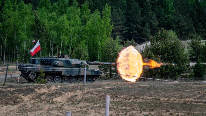 A tracked armoured tank fires its main gun, creating a bright muzzle flash and smoke cloud on a training range; a red-and-white flag is mounted on the turret with dense green forest in the background.