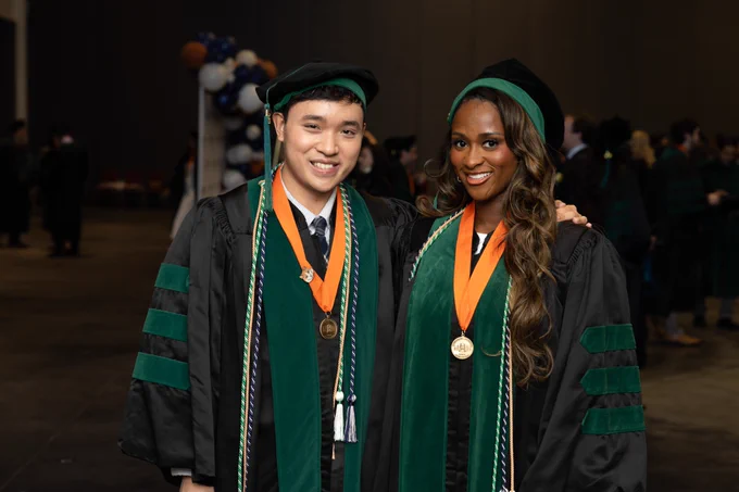 Cody Luong and Ogechukwu Anwaegbu stand together in academic regalia after commencement. Both wear green hoods and orange stoles with honor cords and medallions.