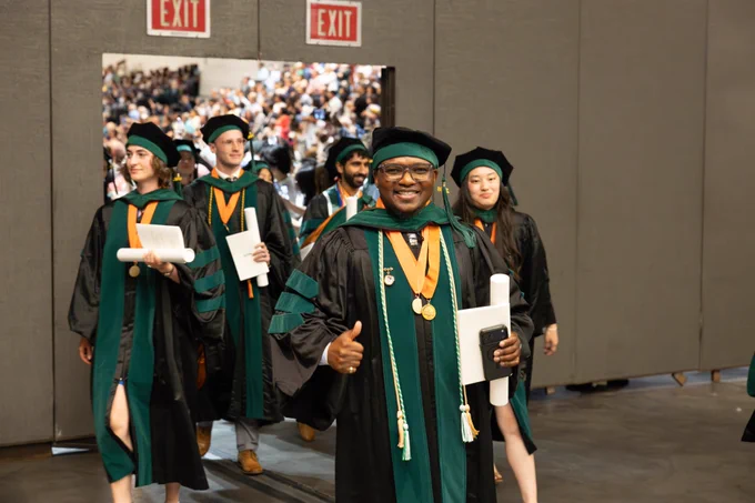 Yves Balikosa smiles and gives a thumbs-up while walking with fellow graduates after commencement. He wears black and green academic regalia and carries his diploma.
