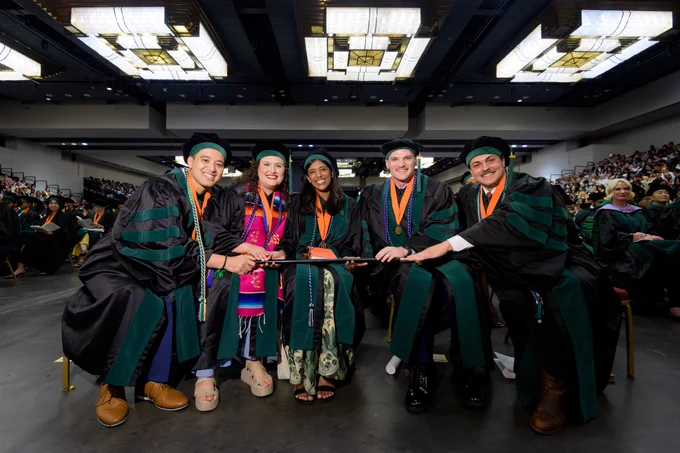 Gold-Headed Cane Award finalists sit together in academic regalia, holding the ceremonial cane across the group. A commencement audience fills the background.